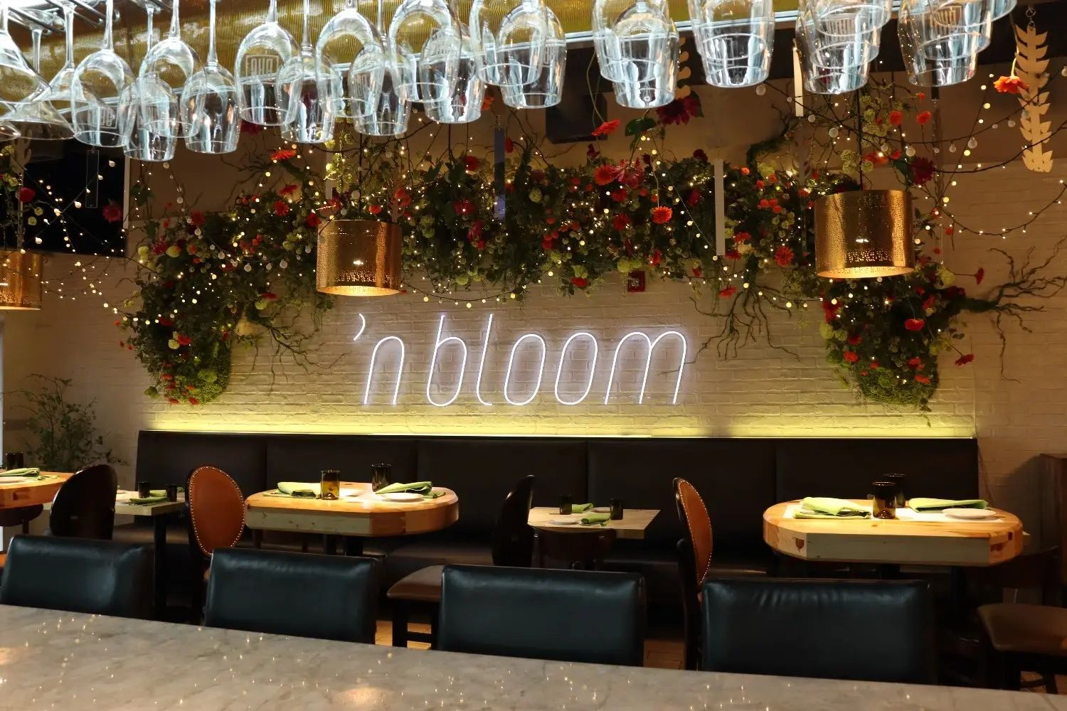 Elegant restaurant dining area featuring a custom white neon sign reading n bloom on a brick wall, surrounded by beautiful floral arrangements, fairy lights, and hanging wine glasses.